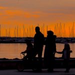 A silhouette of a family walking by the harbor at sunset with boats in the background.